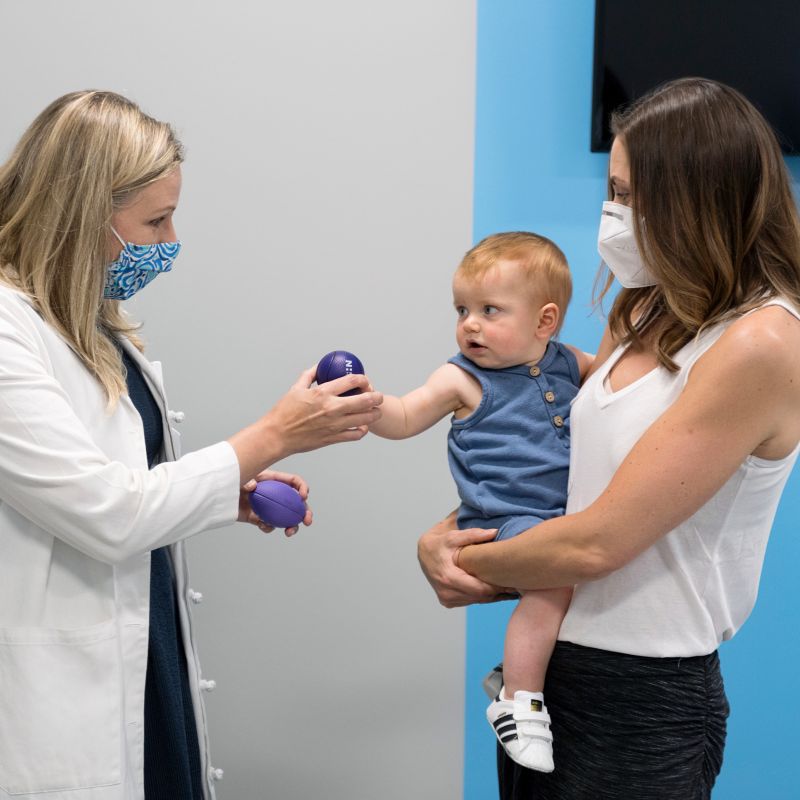 A healthcare provider wearing a mask hands a small purple ball to a baby held by their parent during a pediatric visit.