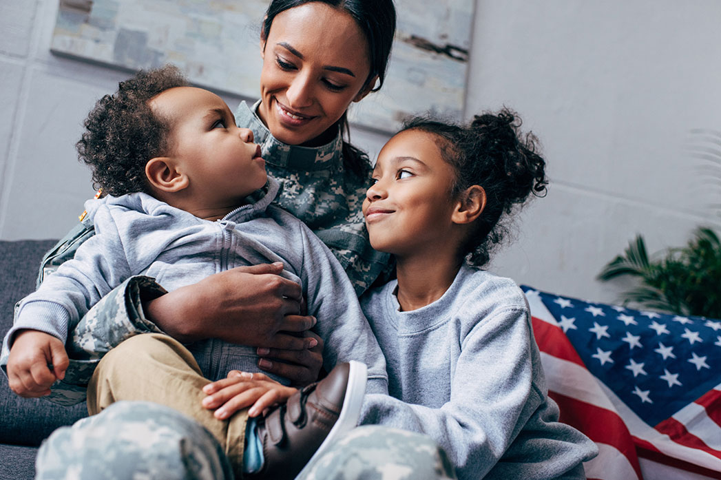 A military service member in uniform sitting with her two young children, smiling together with an American flag in the background.